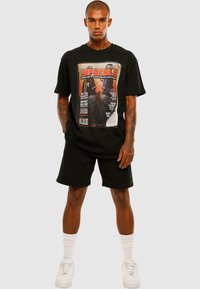 Sort grafisk T-shirt med et magasincover-design, parret med sorte shorts og hvide sokker. Modellen bærer hvide sneakers.