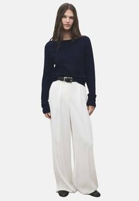 Femme portant un haut noir à manches longues, un pantalon large blanc avec une ceinture noire, et des chaussures noires, se tenant contre un fond uni.