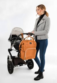 Orange quiltet pusletaske med en frontlomme hænger fra en kørepose. En kvinde i en grå jakke skubber køreposen iført mørke jeans og støvler.