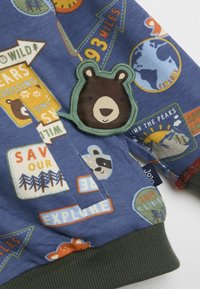 Sinine dressipluus erinevate värviliste loomade ja seikluseteemaliste plaastritega, kaasa arvatud karu plaastriga ja ribaga roheliste varrukatega.