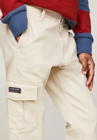 Pantaloni cargo beige con una tasca a toppa, featuring un'etichetta del logo "HILFIGER". Il tessuto appare resistente con una leggera texture.