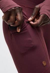 Χέρια που τοποθετούν ένα μαύρο smartphone στην πλαϊνή τσέπη ενός στενού μπορντώ κολάν με ένα μικρό λογότυπο αλεπούς κοντά στο τελείωμα.