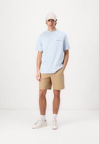 Cămașă de bumbac bleu deschis cu un imprimeu mic pe piept, asortată cu pantaloni scurți bej și adidași albi. Design simplu, croi casual.