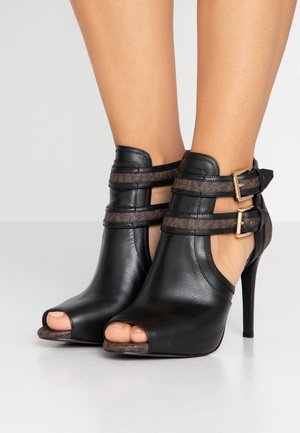 Bottines noires en cuir à talons hauts avec bout ouvert, double brides marron et boucles dorées, portées pieds nus.