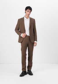 Costum maro, croit dintr-o tesătură texturată, cu rever ascuțit, închidere cu un singur nasture și pantaloni slim-fit. Purtați cu o cămașă albă și pantofi negri.