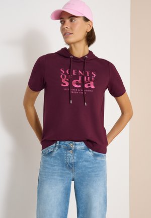Femme portant une casquette rose, un t-shirt à manches courtes avec capuche bordeaux portant le texte "Scents of the Sea", et un jean bleu clair, debout les mains dans le dos.