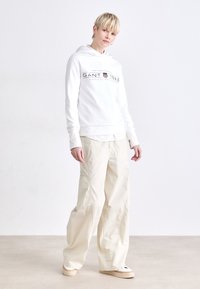 Hvit hettegenser med tekstlogo på forsiden, kombinert med beige vide bukser og hvite joggesko. Enkelt, uformelt design.