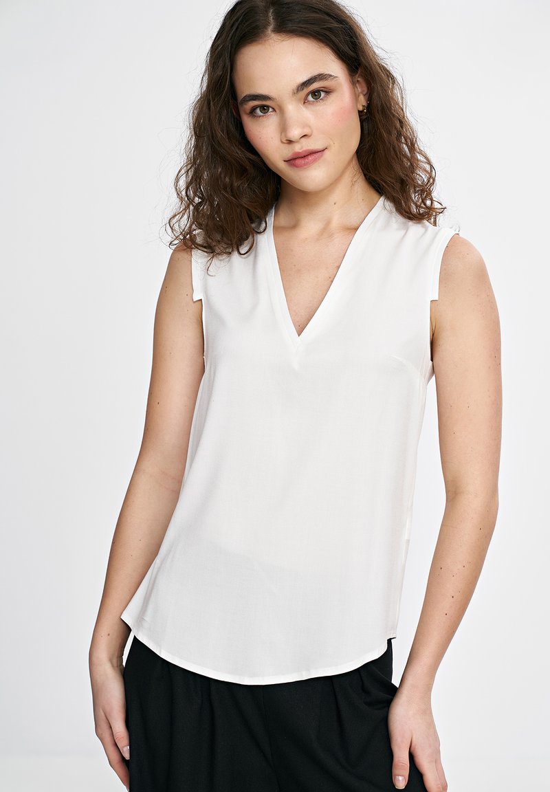 Biała bezrękawnikowa bluzka z dekoltem w serek, o gładkiej fakturze i lekko zaokrąglonym dolnym końcu. Materiał wydaje się lekki i przewiewny.