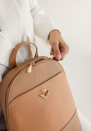 Hnědý kožený batoh se zlatými detaily, se zapínáním na zip a zaobleným designem. Na přední straně je logo a nahoře má držadlo.