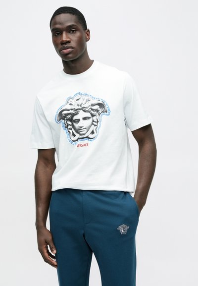 Λευκό κοντομάνικο T-shirt με μεγάλο ασπρόμαυρο γραφικό της Μέδουσας με μπλε περίγραμμα και το κείμενο "VERSACE" σε κόκκινο κάτω από το σχέδιο.