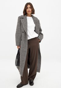 Gray herringbone long coat with large lapels, paired with a white shirt and brown wide-leg trousers, accessorized with a black bag and loafers.
