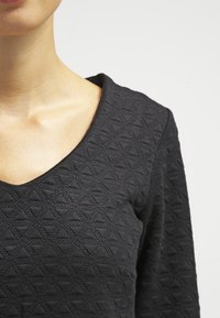 Svart strukturerad topp med V-ringning, med ett geometriskt mönster. Tyget ser mjukt och något elastiskt ut.