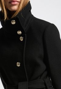 Abrigo de lana negro con cuello alto, que presenta botones dorados y un detalle de cinturón a juego, mostrando una textura en la tela.
