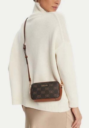 Mujer con un suéter de cuello alto color crema y falda beige, llevando un bolso bandolera marrón con estampado y el logo de "Liu Jo".