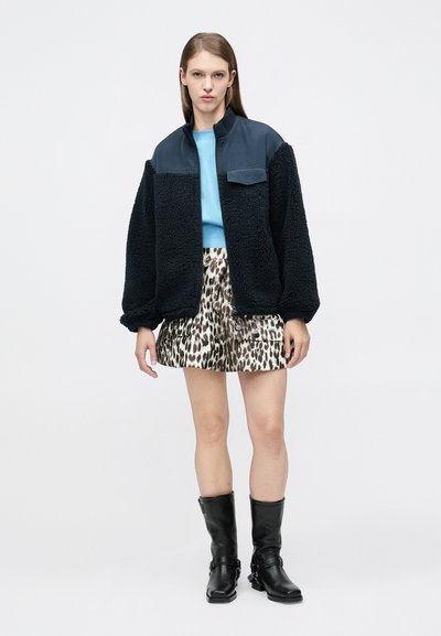 Γυναίκα που φοράει μαύρο φλις σακάκι, ανοιχτό μπλε μπλουζάκι, φούστα με λεοπάρ σχέδιο και μαύρες μπότες μέχρι τα μέσα της γάμπας, στέκεται μπροστά σε λευκό απλό φόντο.