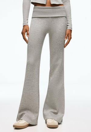 Personne portant un pantalon gris clair à jambes larges et des baskets beiges, debout devant un fond blanc uni.