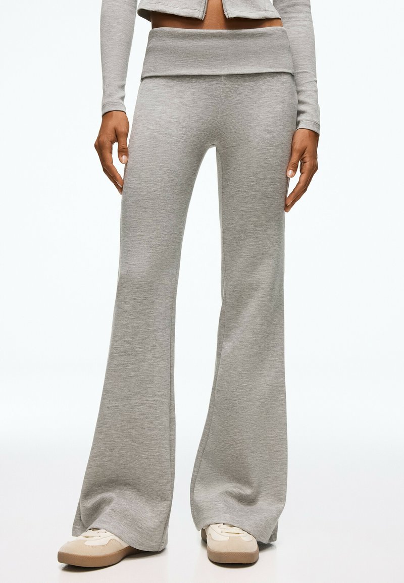 Personne portant un pantalon gris clair à jambes larges et des baskets beiges, debout devant un fond blanc uni.