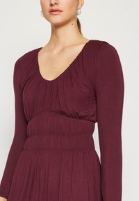 Μακρυμάνικο φόρεμα σε σκούρο μπορντό με στρογγυλή λαιμόκοψη, μαζεμένη φούστα και εφαρμοστό καvasί. Μαλακό, υφασμάτινο ύφασμα με φαρδιά φούστα.