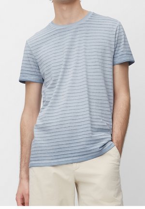 Mężczyzna ubrany w jasnoniebieską, paskowaną koszulkę na krótki rękaw i beżowe krótkie spodenki, z jedną ręką w kieszeni, na jednolitym tle.