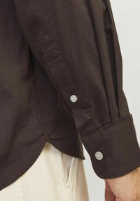 Braunes Langarmshirt mit geknöpften Manschetten, das eine glatte Textur, einen subtilen Glanz und einen abgerundeten Saum mit kontrastierenden Nähten aufweist.