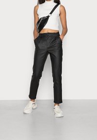 Pantaloni in pelle nera a vita alta con pieghe, abbinati a un top bianco a coste senza maniche e a una borsa a tracolla nera. Le sneakers bianche completano il look.