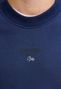 Navy blue sweatshirt with a ribbed neckline featuring printed text "ESS.WRD." and "Studio" in contrasting colors on the front.