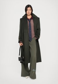 Un manteau en laine vert foncé sur un pull en tricot bordeaux, une cravate à pois navy, un pantalon large vert avec des broderies florales, et un sac à main noir.