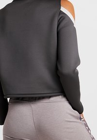 Šedá zkrácená mikina s designem odkrytých ramen, zdobená bílým akcentem na výstřihu. Měkká textura v kombinaci se světle šedými kalhotami.