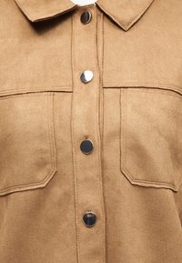 Veste en daim beige devant avec deux poches poitrine et quatre boutons-pression métalliques ronds brillants fermant au centre.