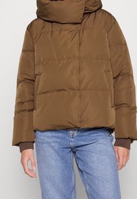 Chaqueta acolchada marrón con un alto cuello doblado, con cierre de cremallera y bolsillos laterales, combinada con unos jeans de mezclilla azul claro.