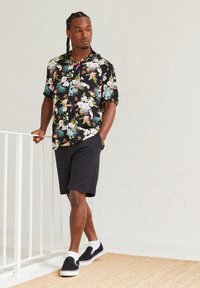 Mann med fletter, iført svart kortermet skjorte med blomster, svarte shorts, hvite sokker og tøysko som lener seg på en hvit rekkverk.