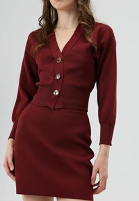 Cardigan en tricot bordeaux avec cinq boutons métalliques et un col en V profond, assorti à une jupe ajustée avec une texture côtelée.