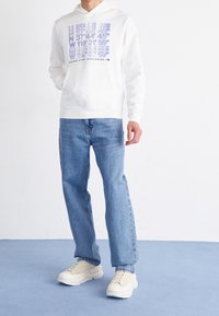 Bílá mikina s kapucí a modrým textem a souřadnicemi, spárovaná se světle modrými džíny a béžovými teniskami. Jednoduchý design, uvolněný střih a jednotná barevná paleta.
