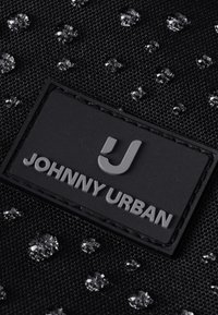 Fekete texturált szövet vízcseppekkel és egy fekete téglalap alakú címkével, amelyen dombornyomott "JOHNNY URBAN" és egy stilizált J logó található.