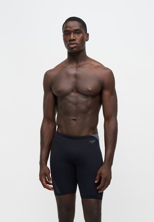 Muskuløs mand, der står fremadvendt iført sorte Speedo svømmejammers mod en ensartet lys baggrund.