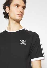 Črna kratkodlaka majica s kratkimi rokavi z belimi detajli, okroglim izrezom in izvezenim logotipom Adidas na levi prsni strani. Mehka tekstura tkanine.
