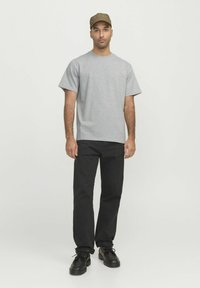 Camiseta de algodón gris de manga corta, combinada con pantalones negros y zapatos negros. El modelo lleva una gorra marrón, de pie sobre un fondo liso.