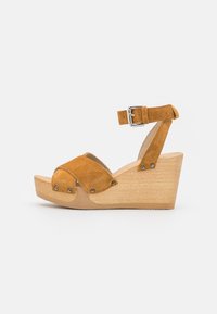 Bruine suede sleehak sandaal met enkelband, met overlappende banden en metalen accenten, geplaatst op een licht houten platform.