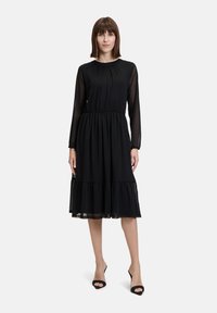 Schwarzes knielanges Kleid mit langen durchsichtigen Ärmeln, gerafftem Taillenbund und schichtsaum. Verfügt über ein dezentes Pünktchenmuster.