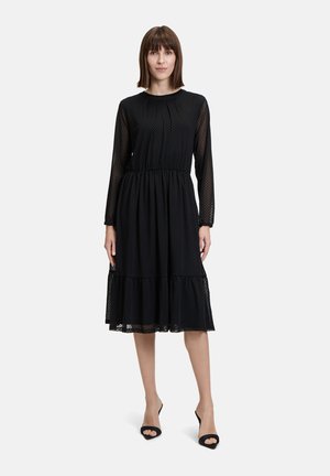 Schwarzes knielanges Kleid mit langen durchsichtigen Ärmeln, gerafftem Taillenbund und schichtsaum. Verfügt über ein dezentes Pünktchenmuster.