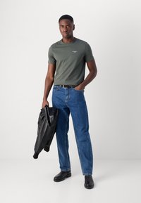 Tricou verde măsliniu pentru bărbați, blugi din denim, jachetă din piele neagră purtată pe braț și bocanci negri până la glezne. Look simplu, casual, cu detalii minime.