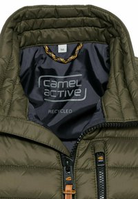 Olivegrüne Steppjacke von innen, mit einem glatten dunkel gefütterten Innenleben, genähten Paneelen, einem Reißverschluss mit Lederzug und dem "Camel Active"-Logo.