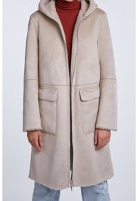 Osoba oblečená v béžovém střiženém kabátu s velkými kapsami, částečně zapnutém, přes hnědý rolák a světlé potrhané džíny.