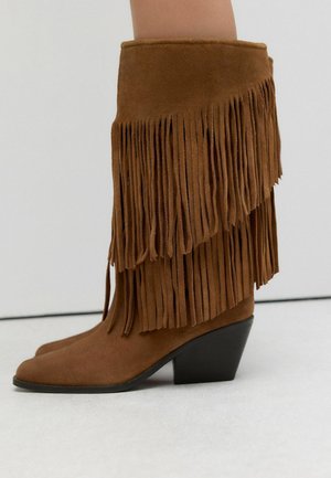 Braune Wildleder-Stiefel bis zur Wade mit geschichteten Fransen und einem hochmodischen schwarzen Wedge-Absatz, die auf einer hellen Oberfläche getragen werden.