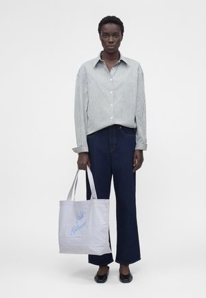 Isik, kes kannab triibulist nööpidega särki ja tumedaid teksaseid, hoides valget kandekotti sinise tekstiga "Café Kitsuné Paris", seisab valgel taustal.