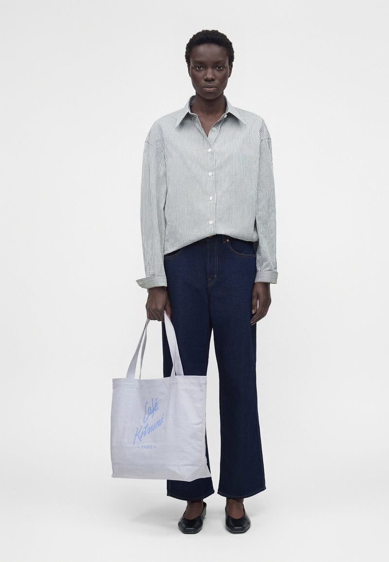 Άτομο με ριγέ πουκάμισο με κουμπιά και σκούρο τζιν, κρατώντας μια λευκή τσάντα tote με μπλε κείμενο "Café Kitsuné Paris", στέκεται σε λευκό φόντο.