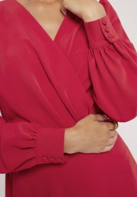 Długa czerwona sukienka o fasonie kopertowym, z bufiastymi rękawami i mankietami na trzy guziki. Gładki materiał i dopasowany krój podkreślają talię.