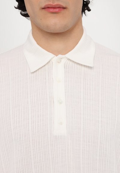 Polo shirt blanc à côtes avec un col doux et un plastron à six boutons comportant de petits boutons irisants. Texture subtile.