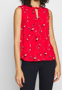 Blouse sans manches rouge avec un décolleté en trou de serrure, présentant un motif floral et des lignes ondulées sur l'ensemble. Fabriquée en tissu léger.