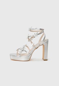 Sølv høyhelede sandaler med sommerfugldetaljer, justerbare stropper og en tykk hæl med platåsåle. Glatt metallisk finish.